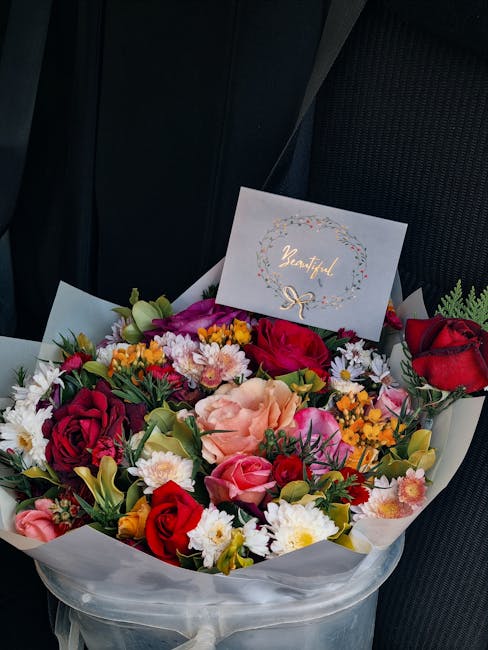 A vibrant floral bouquet arranged with a variety of fresh flowers including red roses, pink roses, peach carnations, white daisies, purple lisianthus, and yellow sprigs, wrapped in white translucent p
