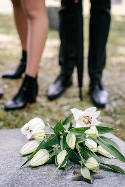A bouquet of white lilies and unopened lily buds with long, glossy green leaves is arranged on a stone surface outdoors. The flowers feature large, trumpet-shaped blooms with prominent stamens, and th