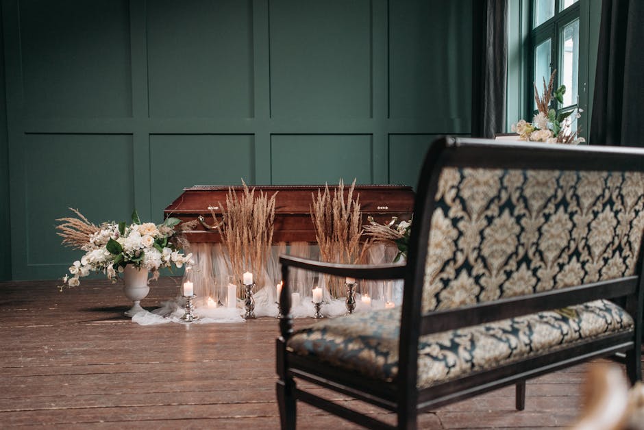 A close-up view of a decorated funeral setting featuring a wooden casket draped with white fabric, surrounded by elegant floral arrangements. To the left, a white ceramic vase holds a bouquet of white