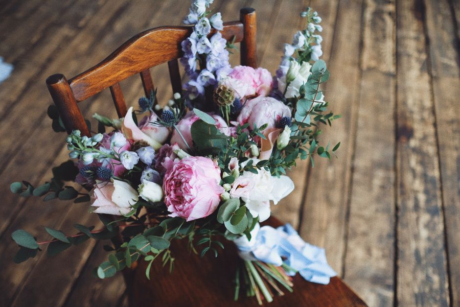 A bouquet arrangement of fresh flowers resting on a wooden chair. The bouquet features large, soft pink peonies and white roses, complemented by sprigs of blue thistle and eucalyptus leaves, creating 