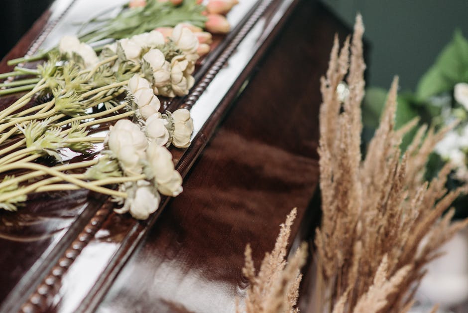A bouquet of white roses with delicate petals, arranged on a polished dark wooden surface next to tall, beige dried ornamental grasses. The roses are fresh, with slightly open blooms and green stems, 