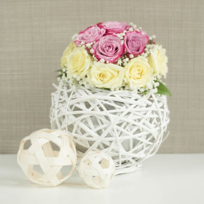 A beautifully styled floral display shows a rose centerpiece resting in a sculptural white wicker vase, captured in soft, even light that feels like a calm afternoon in Kingsbury. About a dozen roses are tightly grouped into a smooth, mound-like shape, each bloom positioned so that the colours flow naturally around the arrangement. The palette is gentle and romantic: half the roses are a pastel pink edging towards lavender or fuchsia, while the other half glow in creamy, buttery yellow that dissolves into ivory at the petal tips. Some buds are still slightly closed, their petals wrapped in a delicate spiral, while others open wider, revealing textured centers that add depth. Interspersed among the roses, delicate sprays of white baby's breath weave through the design, their tiny star-like flowers giving a light, cloud-soft contrast to the more solid rose heads. The flowers are cradled in an eye-catching spherical vase made from thin white branches or wicker, intricately interlaced to form an open-weave cage. Through this airy structure you can glimpse the inner container, a reminder of the skilled craftsmanship behind arrangements created for homes and venues near Kingsbury's Church Lane and surrounding residential streets. The vase stands on a clean white tabletop, whose bright surface enhances the crispness of the whites and subtle tones of the roses. To the left, two natural rattan decorative balls in light wood tones sit on the same table, one slightly larger than the other, echoing the woven textures and adding a curated, contemporary accent. Behind everything, a softly textured beige or light tan wall creates a warm neutral backdrop, ensuring the flowers take center stage. The entire composition feels elegant, tranquil, and ideal for wedding table settings, bridal showers, or sophisticated home décor for gatherings in and around Kingsbury.
