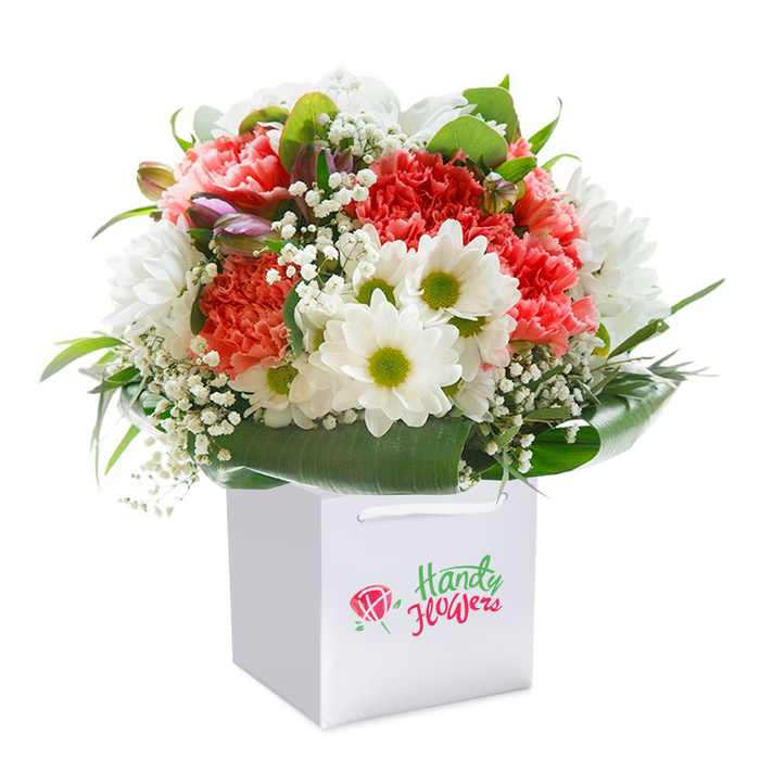 A spectacular floral arrangement, immediately inviting, draws the gaze with its lively interplay of colour and texture-delivered by a skilled Kingsbury florist attuned to the pulse of local occasions and traditions. Predominant coral and peach carnations, their petals artfully ruffled and full, blend seamlessly with a scattering of radiant white, daisy-like chrysanthemums crowned with bright green centers. Each bloom is placed to maximize the interplay of light and shadow, further enriched by lace-like sprays of baby's breath that float between the larger flowers, adding a hint of softness and air. Along the perimeter, several small buds-tinged with a subtle purple blush-offer promise of flowers yet to unfurl, suggesting the bouquet's freshness and attention to detail. Encasing the vivid blossoms, broad, deep green, lacquered leaves form an elegant, sculpted cradle, lending the design a sense of natural grace. The bouquet is presented in a sleek, square white bag with smooth ribbon handles, echoing the clean, contemporary style appreciated by Kingsbury residents along Kingsbury Road and beyond. The white background and professional composition emphasize the bouquet's vibrant hues and impeccable craftsmanship. Every element comes together to convey abundance, sophistication, and heartfelt intent, ensuring this floral gift brings joy-whether for a birthday, anniversary, or just because-throughout Kingsbury. A true testament to the artistry and reliability of local floral delivery, this arrangement exudes both freshness and the spirit of thoughtful giving.