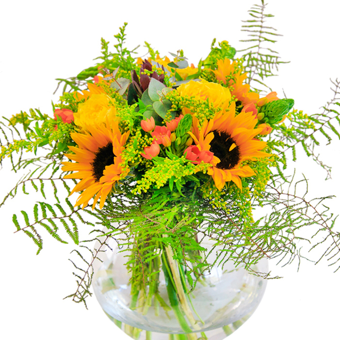 On first impression, this stunning floral display radiates the pure exuberance of a Kingsbury summer day. Clusters of radiant sunflowers and softly spiraled yellow garden roses gather exuberantly, their petals catching the light as if reflecting the golden glow found in local gardens near Roe Green or Salmon Street. The arrangement is intertwined with a variety of deep green leaves and feathery, vine-like ferns, which tumble outward, echoing the exuberance of wild foliage along the Brent Reservoir pathways. Amidst this sea of yellow and green, glossy carmine hypericum berries gleam, bringing energetic pops of red that shine with playful charm. Every stem is elegantly presented in a clear, round glass vase, the water inside refracting the vibrant hues and enhancing the overall feeling of freshness. Subtle eucalyptus leaves thread gently through the bouquet, lending a modern, refreshing whisper and softly masking the warm, honeyed scent that rises from the roses and sunflowers. The tactile contrast between the plushness of the petals, the crisp snap of greens, and the smooth glass suggest a well-crafted arrangement, perfect for moments of happiness: milestone birthdays, anniversaries, or any gathering seeking a bit of sunshine. This bouquet's effortless blend of colour and texture makes it a heartfelt and sophisticated gift, capturing the natural beauty and cheerful outlook synonymous with Kingsbury.