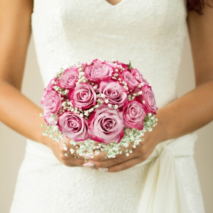 Captured in exquisite detail, this scene showcases a bride, poised amidst Kingsbury's storied charm, her arms embracing a superbly round bouquet centered in the photograph. Approximately a dozen two-tone pink roses anchor the arrangement, their velvety petals radiating from a pale, whisper-soft blush in the heart to edges streaked with saturated fuchsia and magenta. These graceful roses are separated by clusters of frothy white baby's breath, which lend an airy, almost dreamlike textural contrast that harks back to classic bridal traditions appreciated by Kingsbury families for generations. The bride's hands-warm in complexion and tipped with pale, glossed nails-support the bouquet just beneath the intricate, floral lace bodice of her sleeveless gown. A soft sash or tie on her right suggests movement, bringing a relaxed elegance to the portrait, much like the community spirit found along Kingsbury Road. The background fades gently into creamy beiges, drawing the gaze inward to the captivating interplay of blooms and fabric. Every petal, stitch of lace, and swirl of baby's breath tells a story of devotion, artistry, and local wedding heritage, making this image a quintessential representation of Kingsbury bridal style.